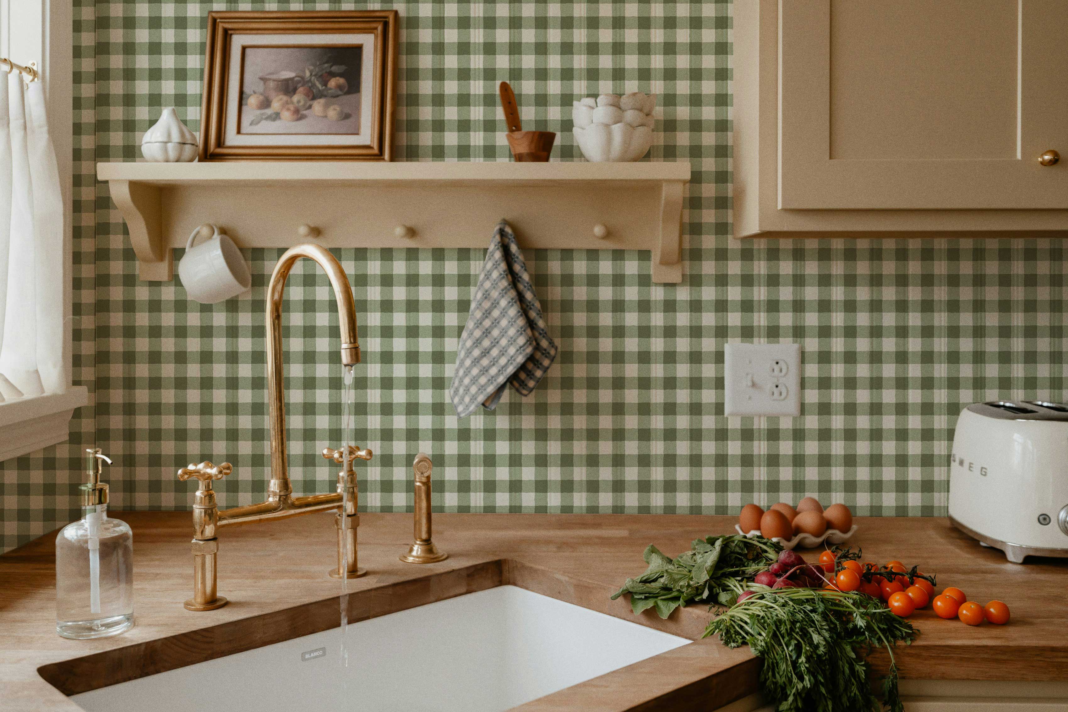 A cozy farmhouse kitchen with green gingham wallpaper, a brass faucet running over a white sink, warm wood countertops, and a small shelf holding vintage decor.