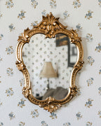 A gold ornate vintage mirror hangs on a wall covered in small blue and muted gold floral wallpaper. The mirror reflects a matching floral-patterned wall, a beige table lamp, and part of a sofa in a softly lit room.