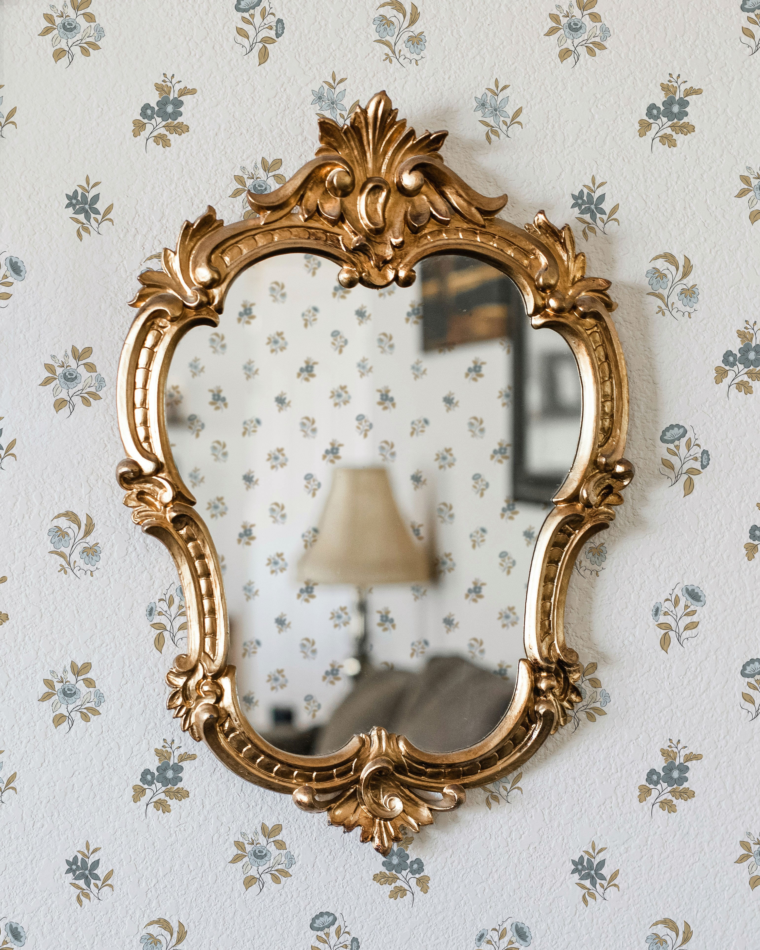 A gold ornate vintage mirror hangs on a wall covered in small blue and muted gold floral wallpaper. The mirror reflects a matching floral-patterned wall, a beige table lamp, and part of a sofa in a softly lit room.