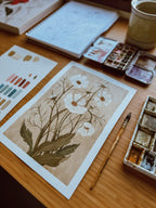 Cosmos watercolor print by Danelys Sidron on studio desk with paint palette, brush, and sketchbook – hand-painted botanical illustration.