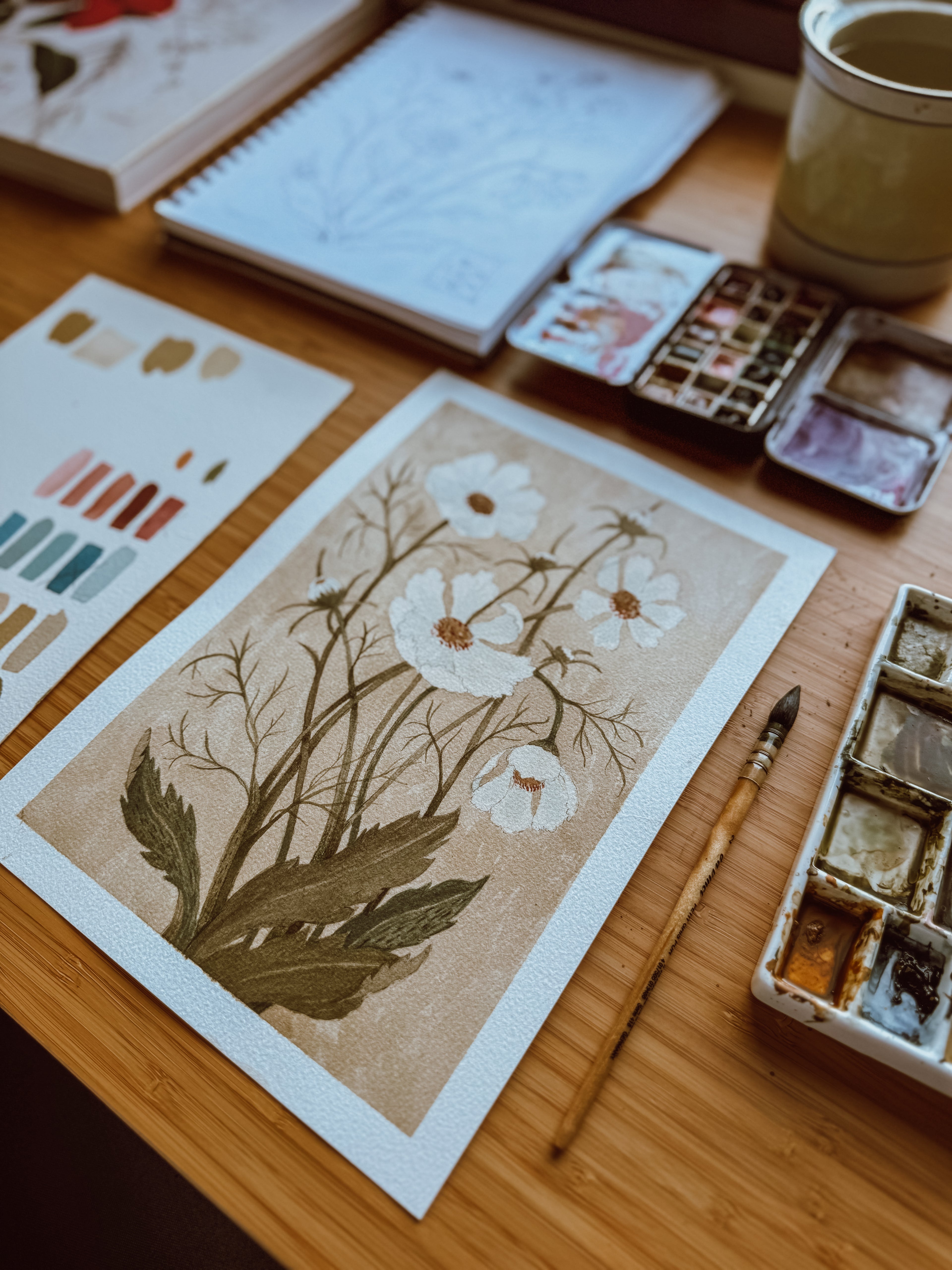 Cosmos watercolor print by Danelys Sidron on studio desk with paint palette, brush, and sketchbook – hand-painted botanical illustration.