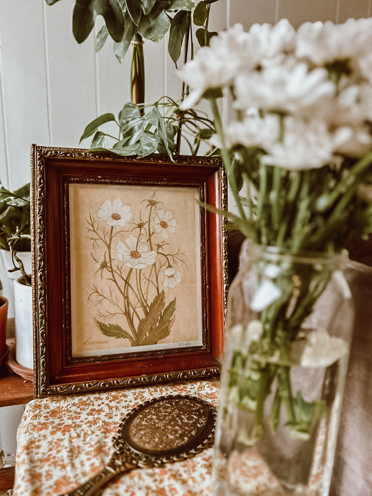 Cosmos watercolor print displayed in an ornate vintage frame beside fresh white flowers – artwork by Danelys Sidron.