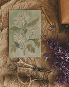 Danelys Sidron Chrysanthemum art card – November birth flower watercolor design photographed on brown paper with scissors and lilac petals, 3.5x5 botanical stationery from The Conservatory Print Club.