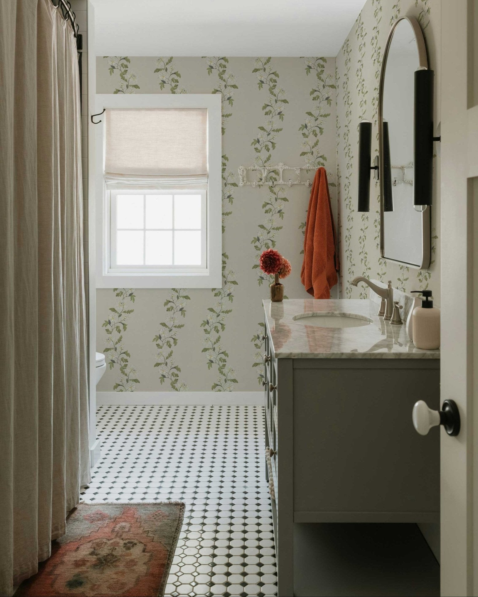 Small bathroom with green floral wallpaper, marble vanity, and black hex tile floor, featuring a window with linen shade and warm vintage accents