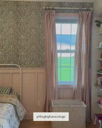 A child's bedroom with an accent wall covered in Roselyn wallpaper in Saybrook Sage, a blue-green botanical pattern. The wall sits above pink painted wainscoting. A white iron bed with a floral quilt is centered in the room, flanked by two windows with pink gingham curtains. A wood dresser and chandelier complete the space.