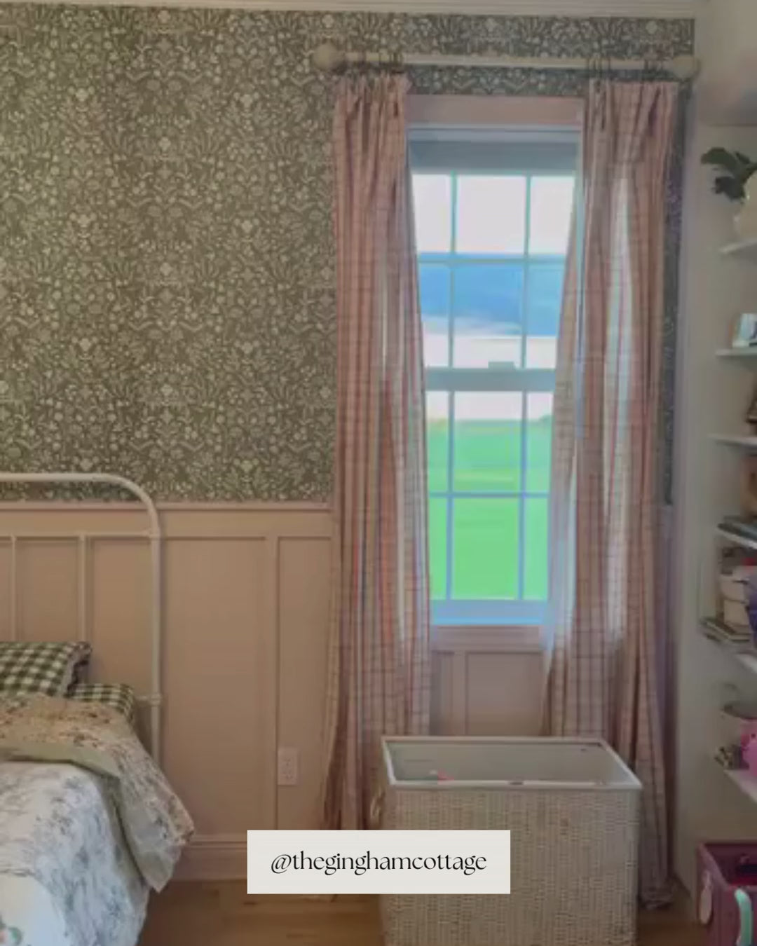 A child's bedroom with an accent wall covered in Roselyn wallpaper in Saybrook Sage, a blue-green botanical pattern. The wall sits above pink painted wainscoting. A white iron bed with a floral quilt is centered in the room, flanked by two windows with pink gingham curtains. A wood dresser and chandelier complete the space.