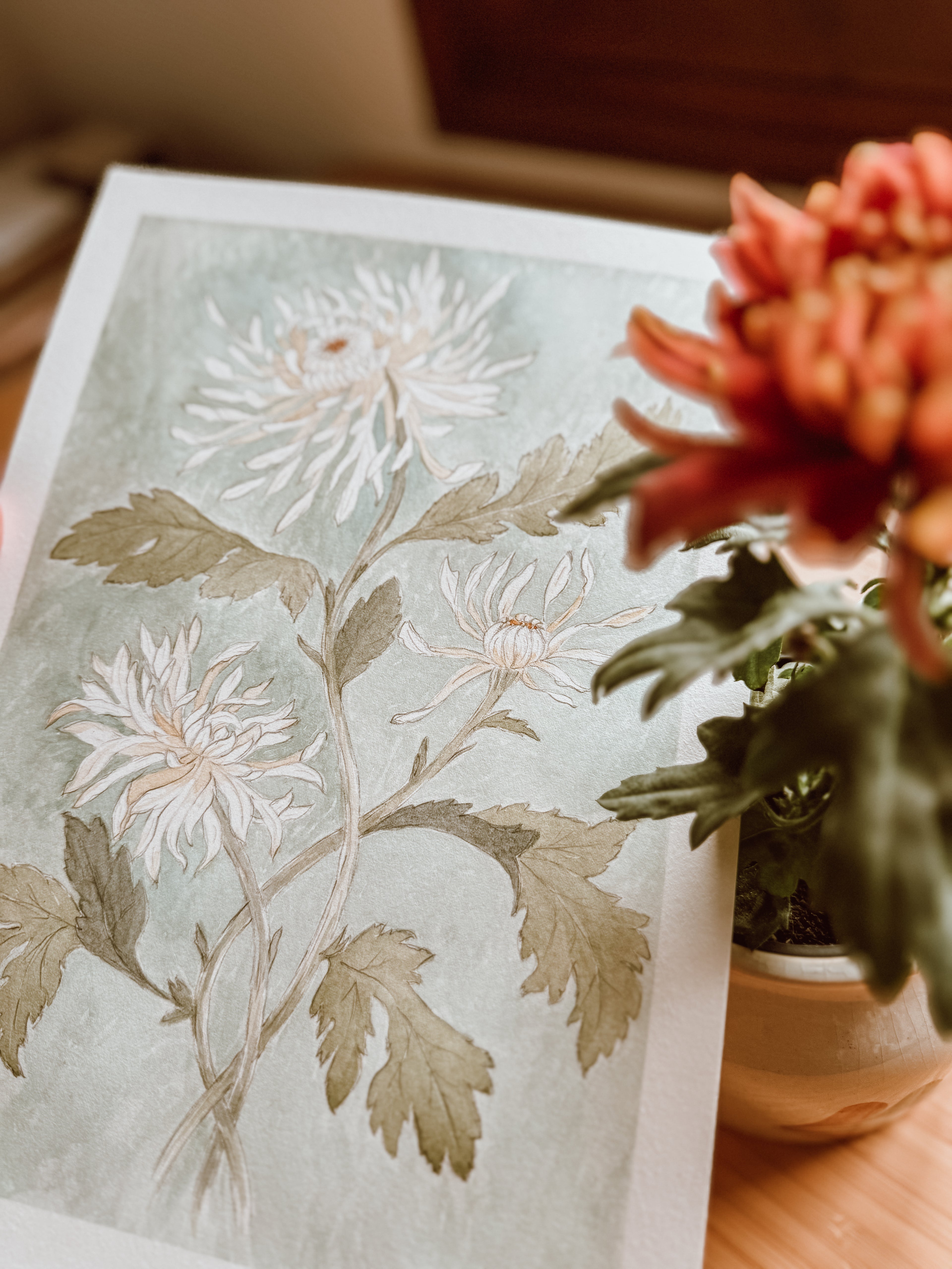 Watercolor chrysanthemum art print styled near a potted flower with soft natural light