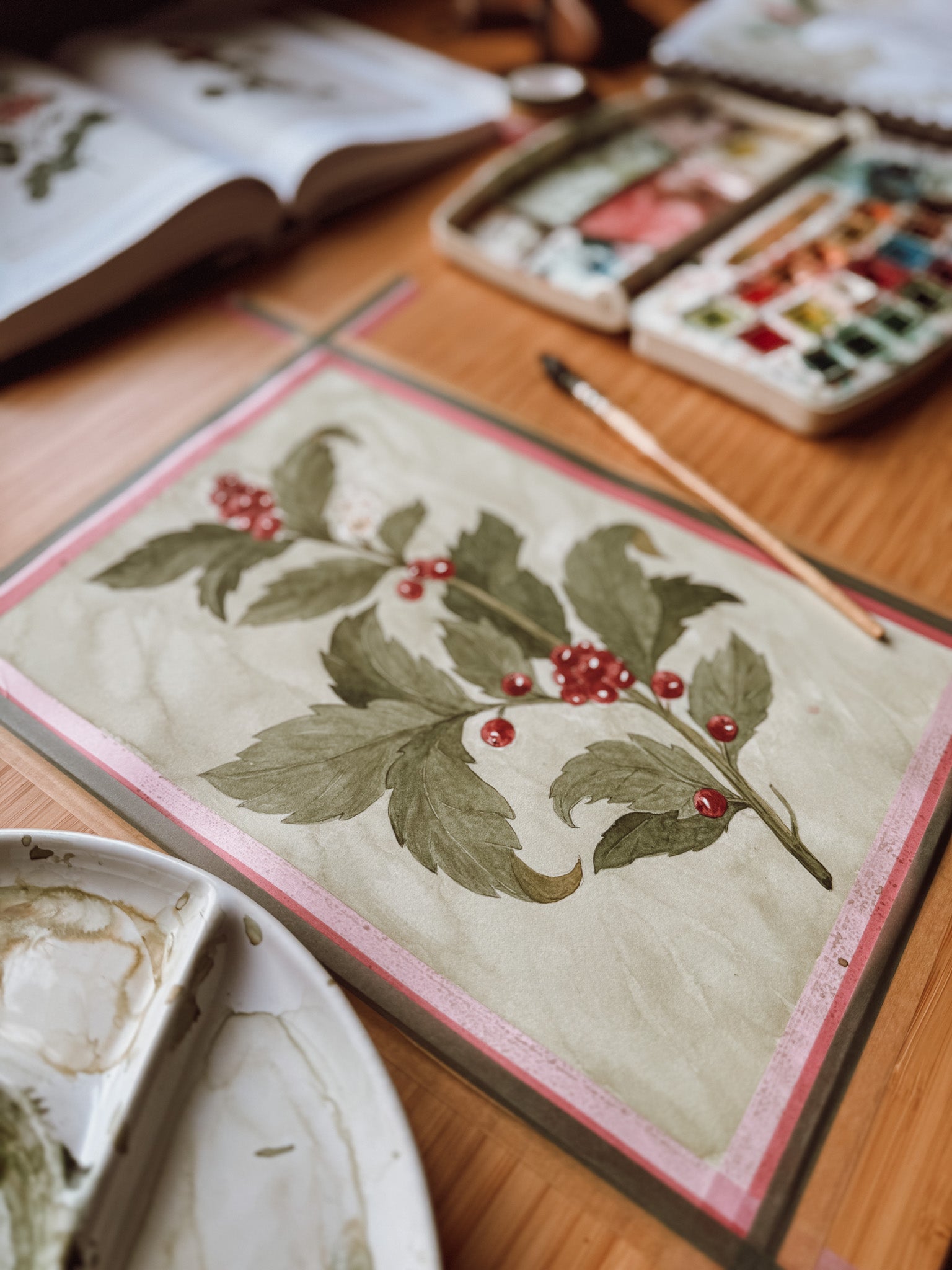 Watercolor botanical painting in progress on a desk, showing holly leaves and red berries with brushes and paints around it