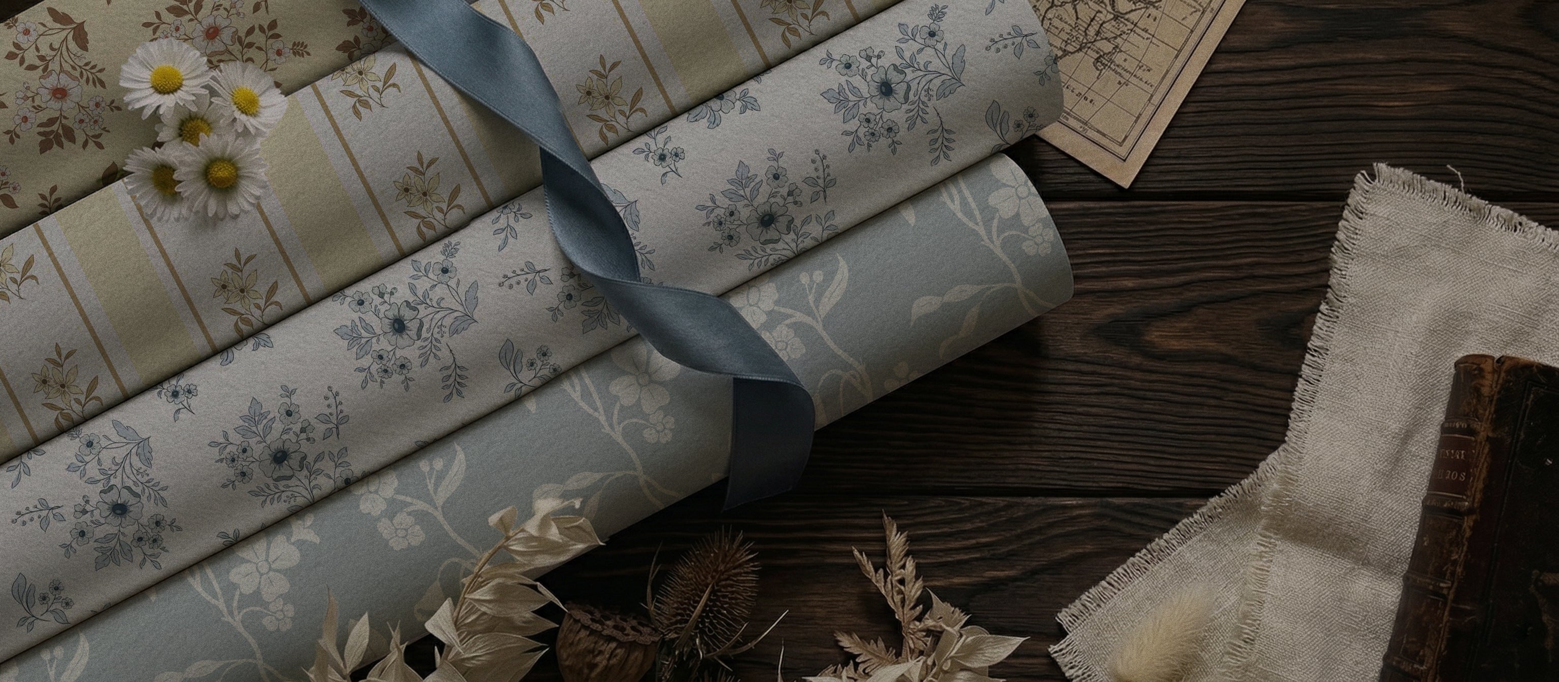 Riverpine Studio botanical and heritage wallpaper rolls in dusty blue and soft cream colorways, styled on dark wood with white daisies and dried botanicals