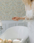 Woman in a white dress sitting in a room with floral wallpaper.