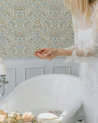Woman in a white dress sitting in a room with floral wallpaper.